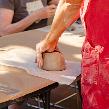 Photograph of a person from below the elbow wearing a red apron. The focus is on their hands as they shape a bowl made of clay at a able covered with a sheet of paper.