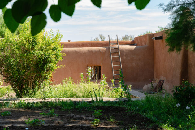 Photograph of an adobe home with a large green garden in front. The wall of the adobe home has an open window and a wooden ladder leans against the wall. Green trees and leaves frame the photograph and above the house is a blue sky.