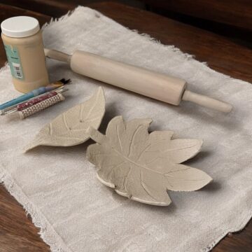 Photograph of two small flat bowls made of clay in the shape of leaves. They rest on a cloth on a wooden table and nearby are small tools