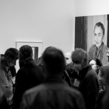 Black and white photograph of a crowd in the foreground and blurry and a photograph of O'Keeffe in the background and in focus.