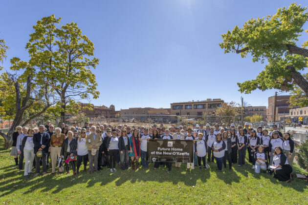 Photograph of a large group of people standing between two trees on a green lawn. They stand behind a banner that says "Future Home of the New O'Keeffe." Behind the group is a construction site. A blue sky in the top half.