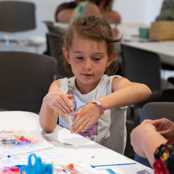 Photograph of a child making a bracelet with colorful beads on a white table. To the right of the frame are a pair of adult hands.