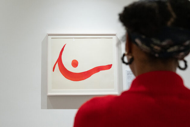 Photograph of a person with dark hair in buns wearing a bright red sweater from behind as they look at a painting by O’Keeffe on a white gallery wall. The painting shows a wave-like line with a dot above in shades of red.