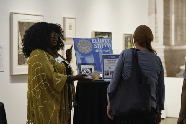 Photograph of a person with dark curly hair standing near a table and showcasing the work of young students to two others in Museum galleries.