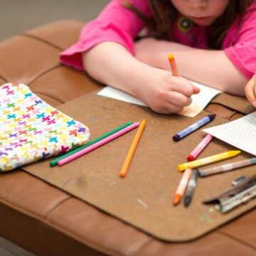 Close-up of a child's hands as they draw with colorful pencils on a leather bench. Their arms are visible in the trop of the frame.