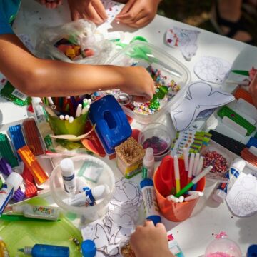 Close-up of colorful art supplies like beads on a table with a group of children's hands using them to make art.