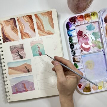 Photograph from above of a person's hands as they paint different small scenes on a notebook with colorful watercolors. To the right is a watercolor palette.