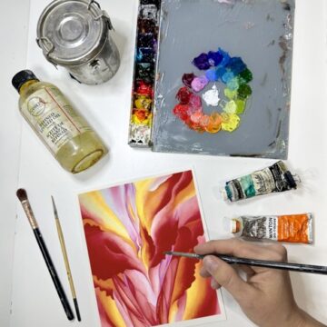 Photograph from above of a person's hands as they paint some abstracted florals in shades of red. Around their canvas are paints, brushes, oil, and a palette.