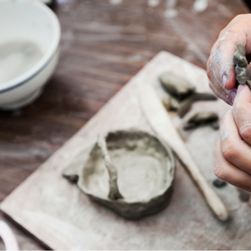 Photograph of hands shaping grey clay above a table where tools and clay are in place to make a bowl.