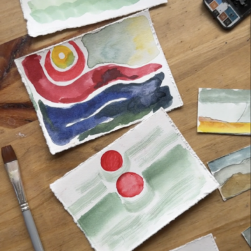 Photograph of three pieces of paper with watercolor paintings of abstract landscapes in shades of red, green, yellow and red. Near the papers are paintbrushes and art supplies. They all sit on a wooden table