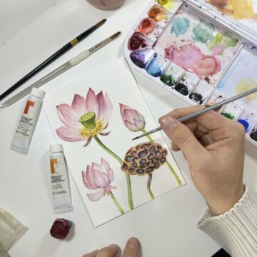 Photograph of an artist's hands from above as they plant several botanical subjects such as a light pink flower. To the right is a watercolor palette.