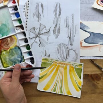 Photograph of an artist's hand as they paint a small canvas in shades of green and yellow. Below their canvas are charcoal sketches of cacti and to the right is a watercolor palette.