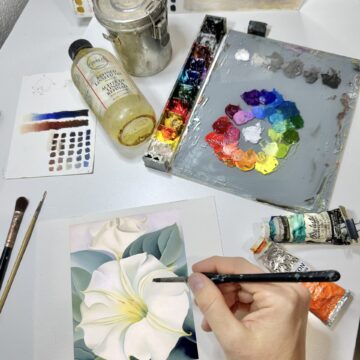 Photograph of an artist's hands as they paint a white flower on a small canvas. Above their hand is an oil pain palette, Lindseed oil, tubes of paint, and other artist supplies.