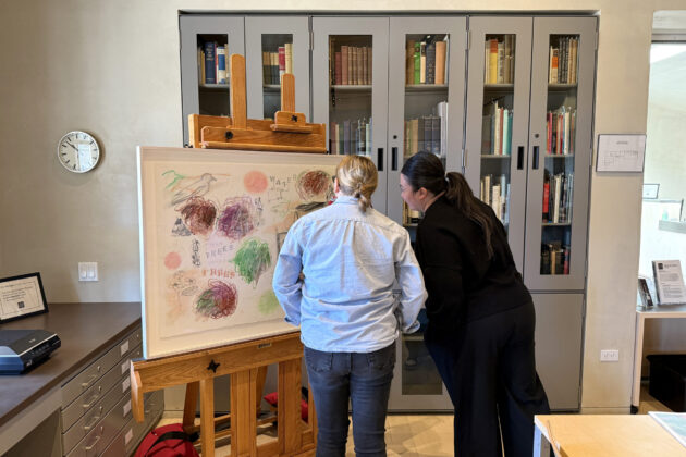 Two people standing looking at a painting on an easel. The painting is a horizontal composition with eight abstract balls of dense scribbled lines in reddish brown, green, white and black pastels. The stenciled words Water, Trees, and Peyote float within the composition surrounded by small singular portraits of five Native Americans along with outlines of buildings, trucks, and ravens or crows.