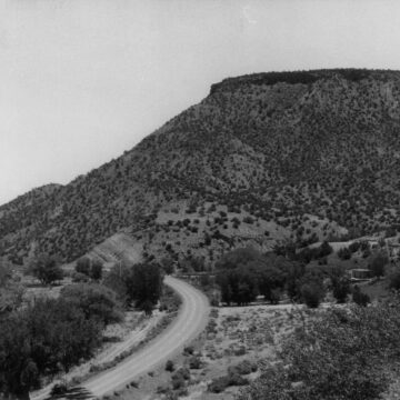 A black and white photograph of the view from O'Keeffe's Home and Studio. The view is a winding road that disappears into the New Mexico hills. A large cliff with scrubby desert tree emerges from the background.