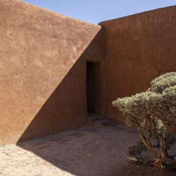 This photograph features a corner of the outside patio at Georgia O'Keeffe's Abiquiu Home and Studio. A shadow darkens half the wall in a diagonal. The brown adobe walls are contrasted by a bright blue sky.
