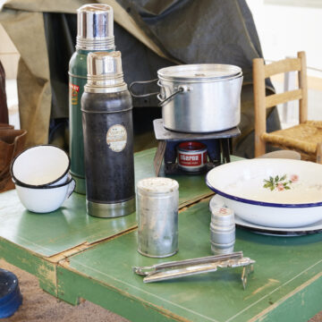On a green worn table sits a white metal soup bowl with a small floral decoration atop a matching plate accompanied by two matching stacked coffee cups. Two vintage thermoses , and a metal camping pot also sit on the table. A small chair is in the background along with a vintage lantern, leather boots, a basket and a tent made of a tarp.